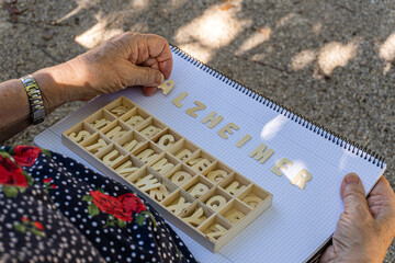 Obraz premium Hands of an elderly woman with Alzheimer's, forming the word Alzheimer with wooden letters on a notebook. Outside a park.