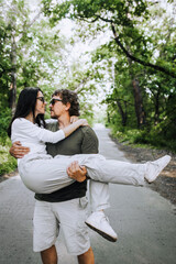 Fototapeta premium A bearded long-haired stylish man holds his beloved hippie brunette woman in sunglasses in his arms and circles her in nature in the park. Portrait, photo of happy and smiling newlyweds.