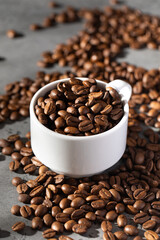 Roasted coffee beans in a white mug, coffee beans scattered around. 