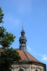 Historische Gebäude mit einem Türmchen aus Schiefer und einem roten Dach. Die Kirche wird im Vordergrund von einem Baum verdeckt und steht vor einem blauen Himmel.