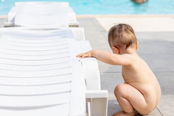 cute little baby boy is playing in pool area,sun lounger,summer sunny day.naked toddler with no...