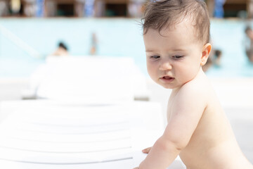 cute little baby boy is playing in pool area,sun lounger,summer sunny day.naked toddler with no...