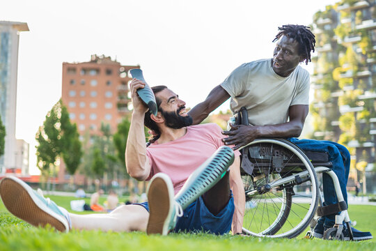 Portrait of two disabled adults. Hispanic man with prosthetic leg joking and showing a piece of prothesis his friend while chatting on the grass