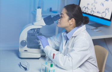 Concentrated young Indian female lab technician in white coat sitting at desk and working with microscope in modern laboratory