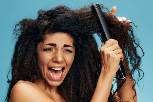 Screaming pretty Latin woman suffer with curly dry damaged hair problems using a hair straightener, posing isolated on blue wall background. Hair routine concept, haircare, hair ironing, hairdressing