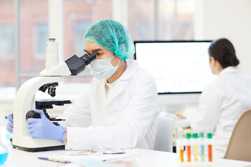 Serious concentrated young Asian male scientist in sterile cap and mask sitting at table and analyzing test sample through microscope in laboratory