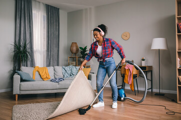 Happy young african american woman in headphones vacuuming with vacuum cleaner in living room - obrazy, fototapety, plakaty