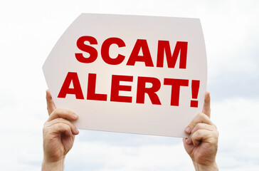 Against the background of the sky in the hands of a man is a sign with the inscription - Scam Alert