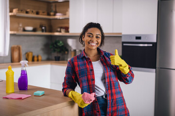 Cheerful millennial african american female in rubber gloves with rag and cleaning supplies showing...