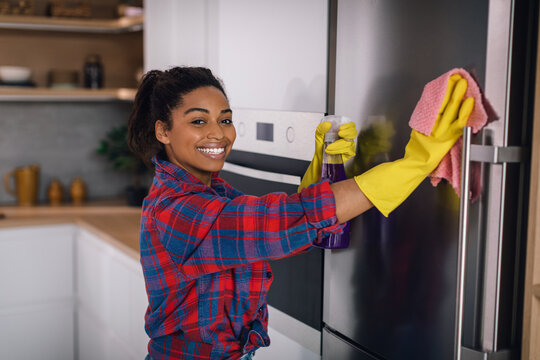 Happy young african american woman in rubber gloves wipes refrigerator with spray and sponge
