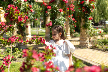 Summer portrait of pretty little girl wearing white dress, posing in red rose garden smelling flowers. Happy smiling girl