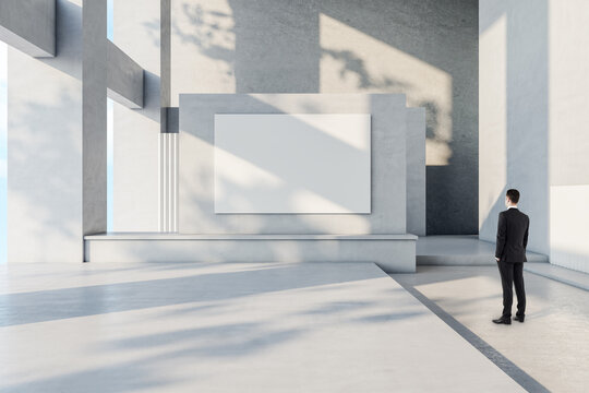 Businessman in suit back view looking at blank white banner with place for your logo or text on light grey wall in sunlit spacious hall with concrete design, mock up - Powered by Adobe
