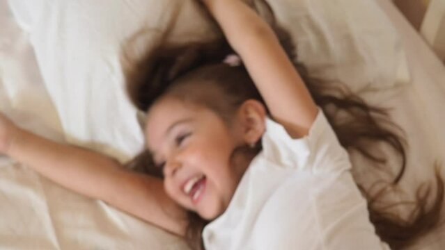 Little girl with tablet and headphones on the bed at home. Child girl listening to music or watching cartoons, chatting with friends or family before bed