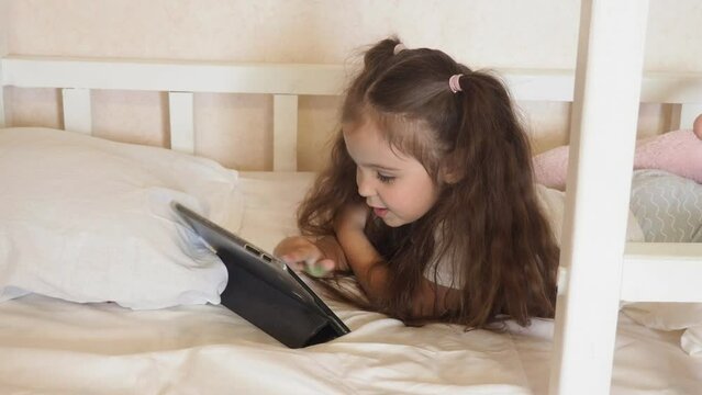 Little girl with tablet or phones on the bed at home. Child girl listening to music or watching cartoons, chatting with friends or family before bed