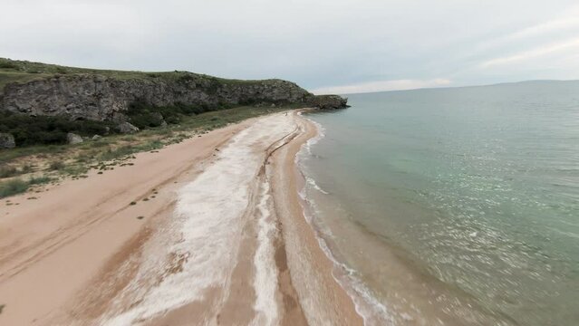  A long empty beach with the sea. Shot . Sand next to small waves and large stone mountains next to standing in a row and blue sky.