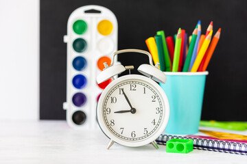 Time to return back to school. Bright colourful green yellow, blue stationery, paints, pencils, markers, brushes. black board on background. White alarm clock. Copy space for text. Autumn, September