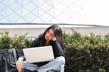 Young bored girl student sitting on city street bench using laptop computer studying