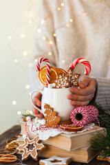 Hot winter drink in a white mug: cozy home composition with homemade gingerbread cookies, candy cane, fir tree branch. Wooden background, christmas lights and candles
