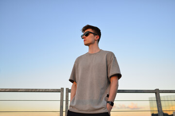 Fototapeta premium Portrait of handsome confident stylish hipster man in sunglasses standing on porch looking at sunset, calm relaxing moment, summer vacation concept