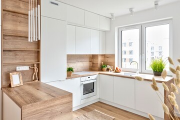 Modern interior of kitchen with white furniture, wooden counter and wooden floor. Kitchen sink with faucet near window and stylish design in new apartment. - obrazy, fototapety, plakaty