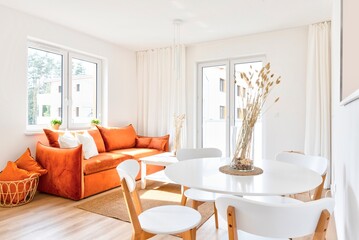 Comfortable red sofa with pillows and stylish round table and wooden chairs in modern living room with window and white wall. Luxurious interior of lounge with dining room. New apartment in hotel.