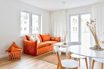 Modern interior of living room and dining room in open space. Round table with chairs and stylish red sofa with pillows in cozy room with window and wooden floor. New apartment in hotel.