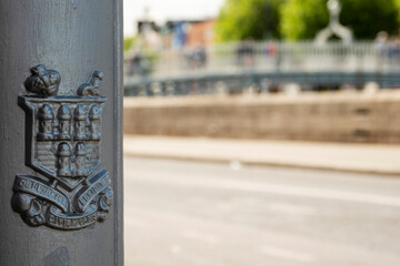 Coat of arms symbol of Dublin on a metal lamp post in focus. Ha'penny Bridge out of focus in the background. City life. Popular tourist area.