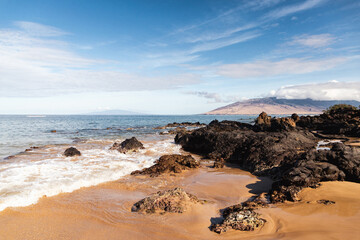 A rocky beach in Maui Hawaii with sand, waves and a mountain in the distance