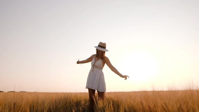 Happy free young woman dancing and spinning around in slow motion across field, touching ears of wheat with her hand. Wheat field on sunset background