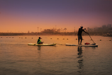 Naklejka premium father and son surfer rides by paddle board (S.U.P.) in the ocean against the background of costa verde (lima,peru) on sunset