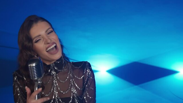 Beautiful Caucasian girl singing into a microphone on a blue background. European woman singer on the spotlight. Shooting a video. Brunette in a shiny evening dress. Music band.