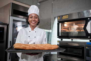 Portrait of African American female chef in white cooking uniform looking at camera with cheerful smile and proud with tray of baguette in kitchen, pastry foods professional, fresh bakery occupation.