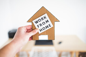 Fototapeta na wymiar work from home sign being hold in front of out of focus home office desk setup, digital nomads working remotely or wfh days during lockdowns