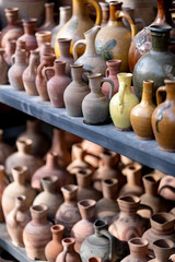 Fototapeta na wymiar Many unique handmade colorful clay jugs and vases of various shapes on shelf, blooming flower plants in local market in sunlight, selective soft focus. Lot of handicraft ceramic pots, top view.