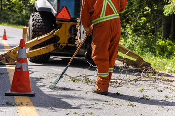 Closeup view as a worker is seen wearing high visibility clothes, using a rake to clear tree branches and leaves from road with tractor in background.