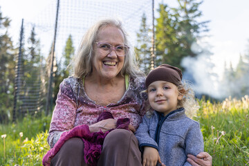 Closeup joyful portrait of elderly smiling grandmother with arm around cute four year old grandson sitting outdoors in nature with blurry background. - Powered by Adobe