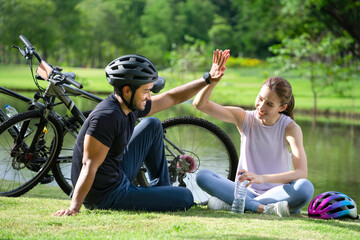 young asian couple hands high five together while resting beside the pond in the park, happy family sitting in the sun after ride bicycle in the morning.