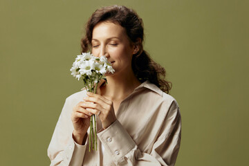 Smiling cute curly beautiful female in linen casual shirt hold chamomiles flowers near face enjoying smell posing isolated on over olive green pastel background. Nature is beauty concept. Copy space