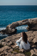 Relaxed woman from behind sit at the edge and looking at sea cave arch bridge