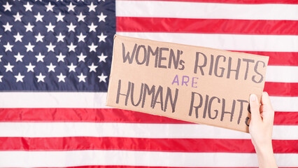 Woman is holding a sign Womens Rights Are Human Rights US flag on background. Protest against anti...