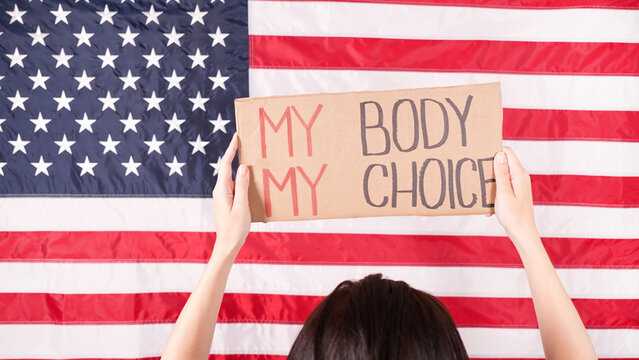 Young woman protester holds cardboard with My Body My Choice sign against USA flag on background. Girl protesting against anti-abortion laws. Feminist power. Equal opportunity Womens rights reedom.