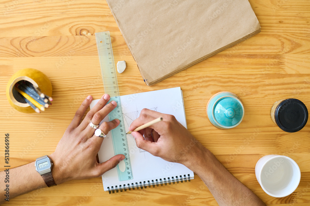 Poster above shot of hands of multi-ethnic guy making sketch of new item of earthenware while sitting by wo