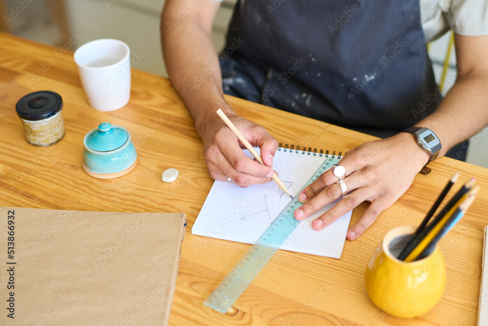 Poster hands of young man in apron sitting by wooden table and making sketch with pencil and ruler of creat