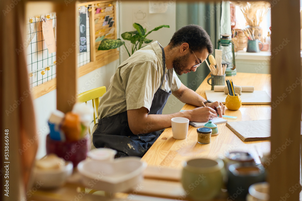 Aufkleber Young creative man bending over workplace while making sketch in notepad in spacious studio or workshop with displays for new items