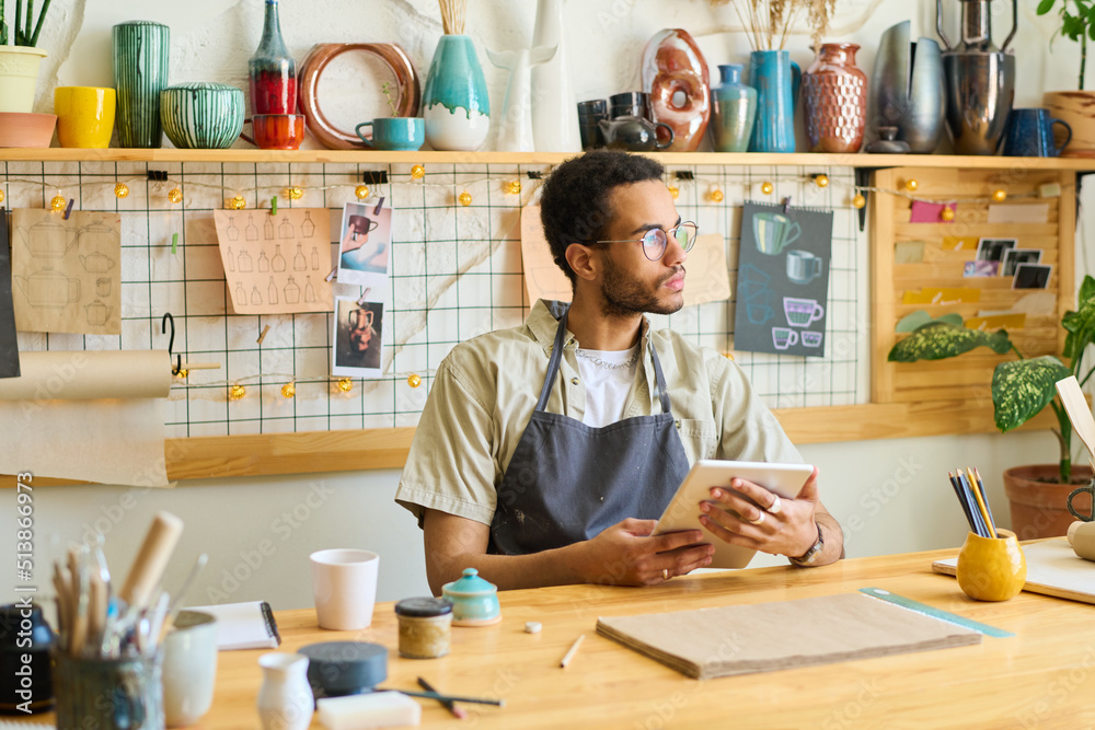 Poster young pensive black man in workwear using digital tablet while sitting by table in workshop and scro