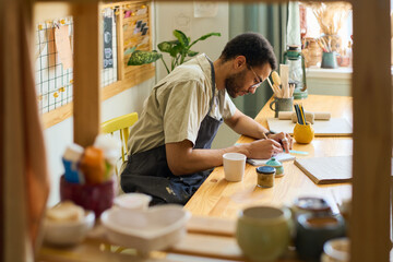 Young creative man bending over workplace while making sketch in notepad in spacious studio or...
