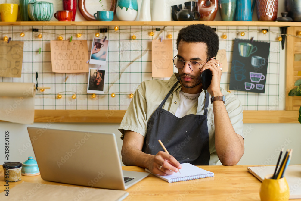 Poster young serious clerk of small earthenware shop talking to client by mobile phone while sitting by tab