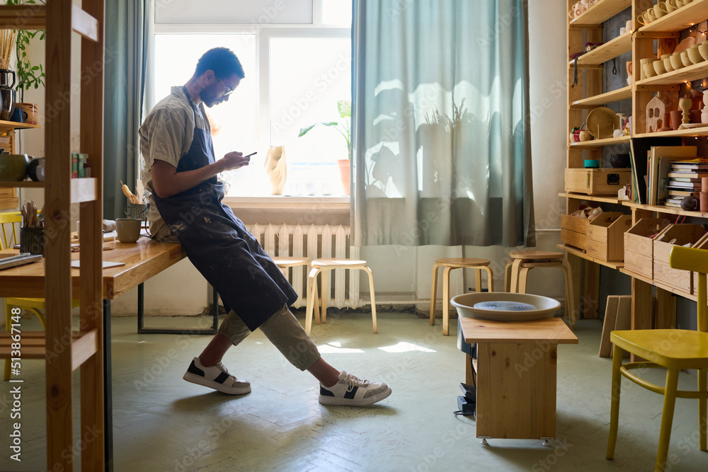 Poster side view of young male artisan in workwear scrolling in smartphone by workplace while looking throu