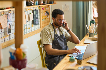Young male clerk in apron calling clients and looking through online orders while sitting by...
