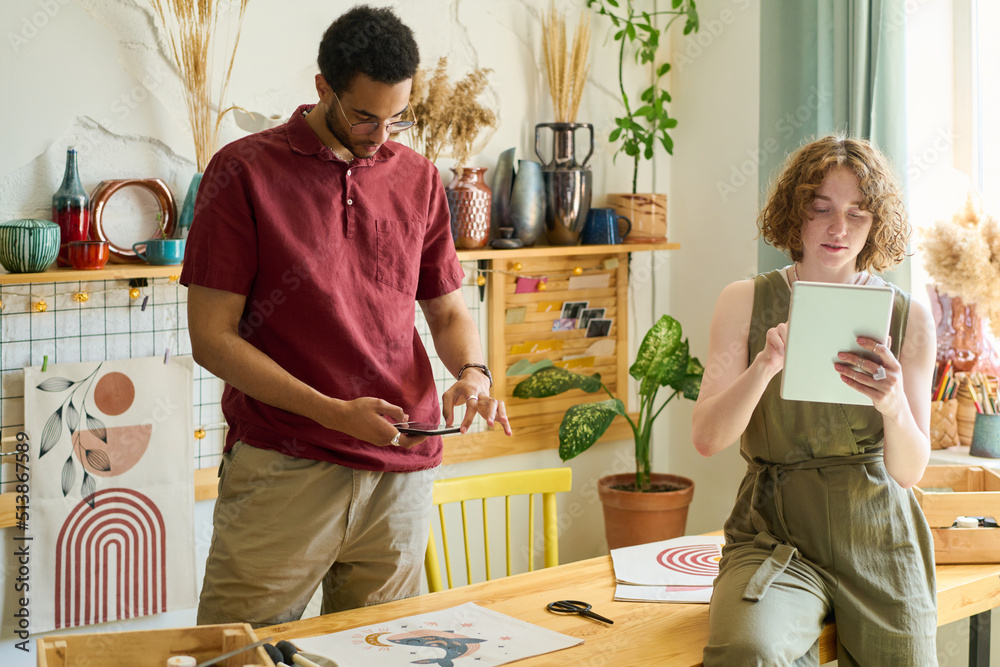 Poster Young female with digital tablet networking while black man with smartphone taking photo of drawing on table in studio